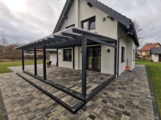 Nowoczesna, grafitowa pergola z zadaszeniem przy białym domu. Widoczna kostka brukowa i fragment ogrodu. Minimalistyczny design, stonowana kolorystyka.