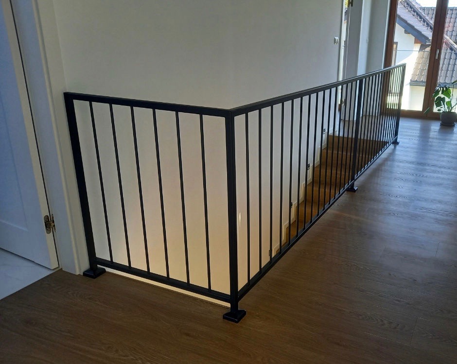 Minimalistyczna balustrada schodowa z czarnych, pionowych prętów i białego wypełnienia, zamontowana w nowoczesnym wnętrzu z drewnianą podłogą i widokiem na ogród.