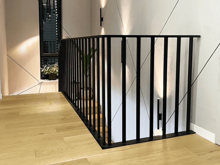 Nowoczesna, czarna balustrada metalowa z pionowymi szczeblami, zamontowana wewnątrz budynku. Widoczne oświetlenie ścienne i drewniana podłoga.