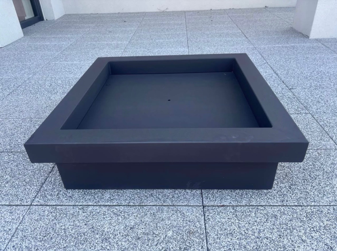 Kwadratowa, metalowa konstrukcja w kolorze grafitowym, prawdopodobnie donica lub obudowa paleniska, ustawiona na szarym, płytkowym podłożu. Minimalistyczny design.
