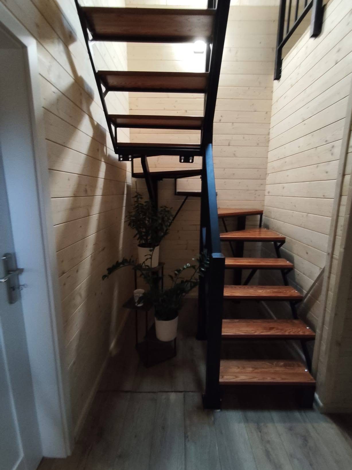 Drewniane schody z metalową konstrukcją w jasnym wnętrzu wykończonym boazerią. Obok schodów widoczne rośliny doniczkowe na małym stoliku.