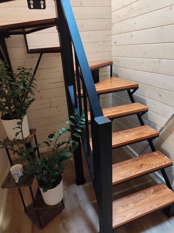 Drewniane schody z czarną metalową balustradą w jasnym wnętrzu z boazerią. Na podeście rośliny doniczkowe. Widok z dołu, ukazujący solidną konstrukcję i estetyczne wykończenie.