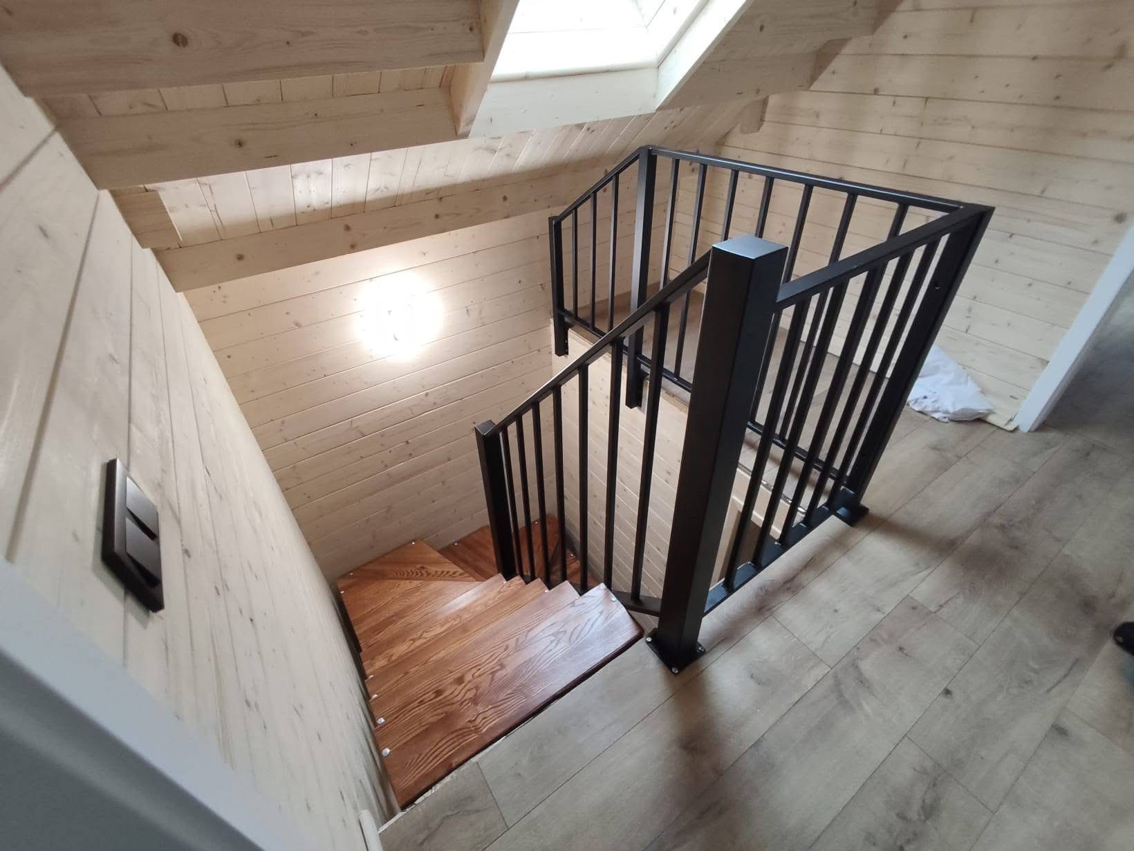 Drewniane schody z czarną, metalową balustradą w jasnym wnętrzu z boazerią. Widok z góry, podkreślający solidne wykonanie i nowoczesny design.