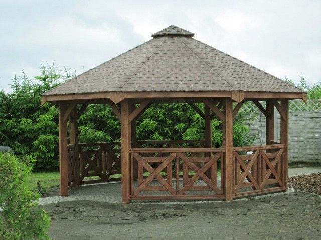 Drewniana altana ogrodowa z brązowym dachem, otoczona zielenią. Konstrukcja z krzyżowymi zdobieniami, widoczna na tle drzew i fragmentu ogrodzenia.