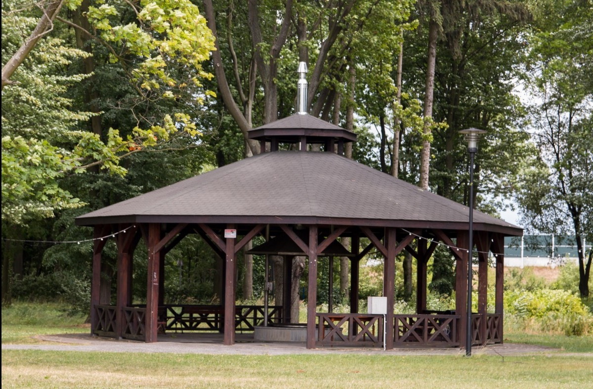 Drewniana altana ogrodowa z grillem pośrodku, otoczona zielenią. Konstrukcja z ciemnego drewna, zadaszona, z ozdobnymi barierkami. Widok z zewnątrz, w tle drzewa i trawnik.