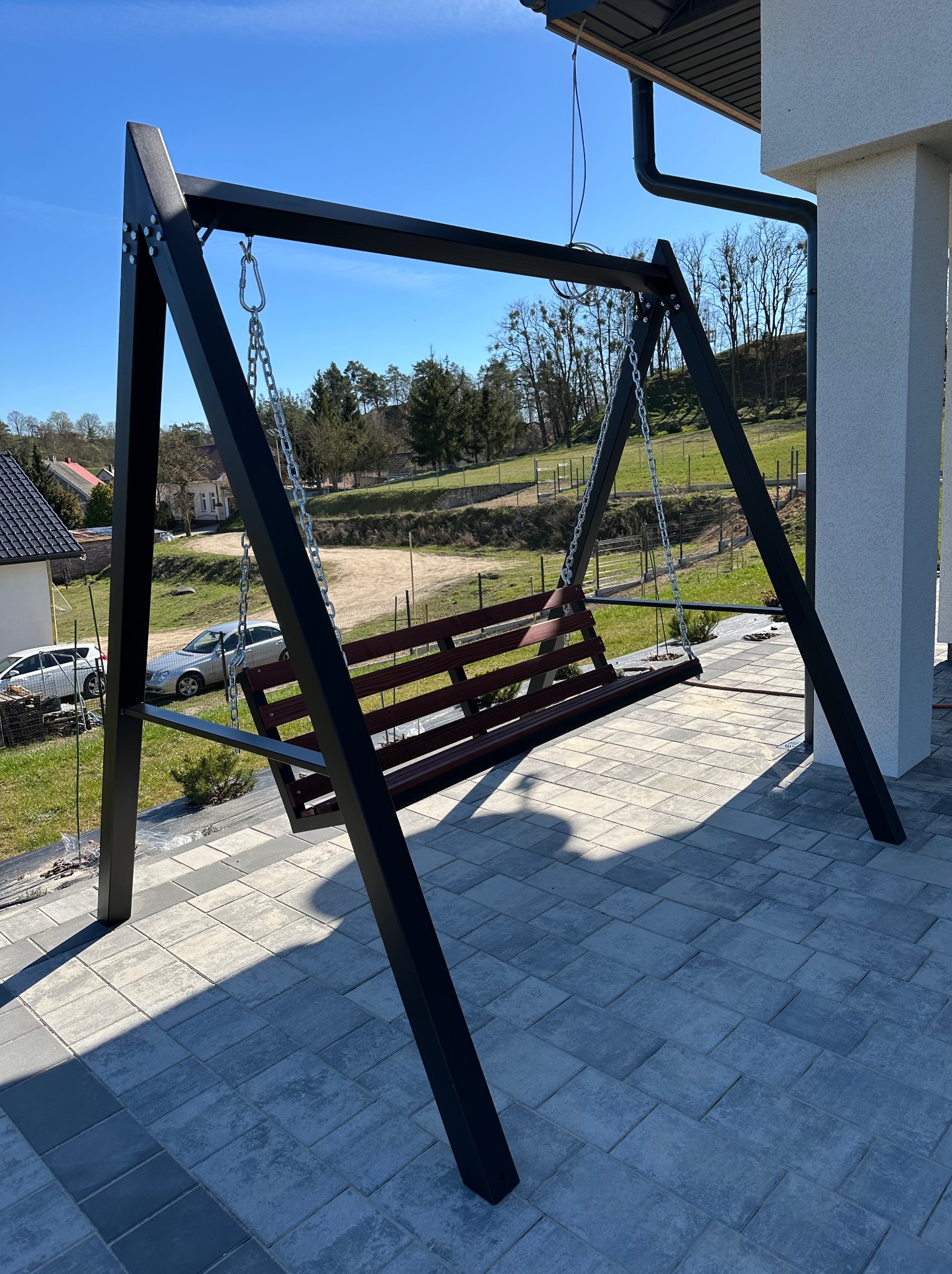 Metalowa huśtawka ogrodowa z drewnianym siedziskiem, zawieszona na łańcuchach, na tle kostki brukowej i zieleni. Konstrukcja pomalowana na czarno, widoczne detale mocowań.