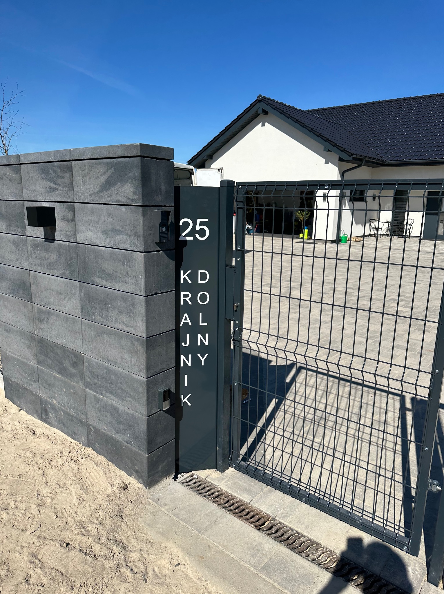 Nowoczesne ogrodzenie panelowe z szarego betonu i metalowej siatki, z numerem domu '25' i nazwą ulicy 'Królnik' na tle błękitnego nieba i domu.