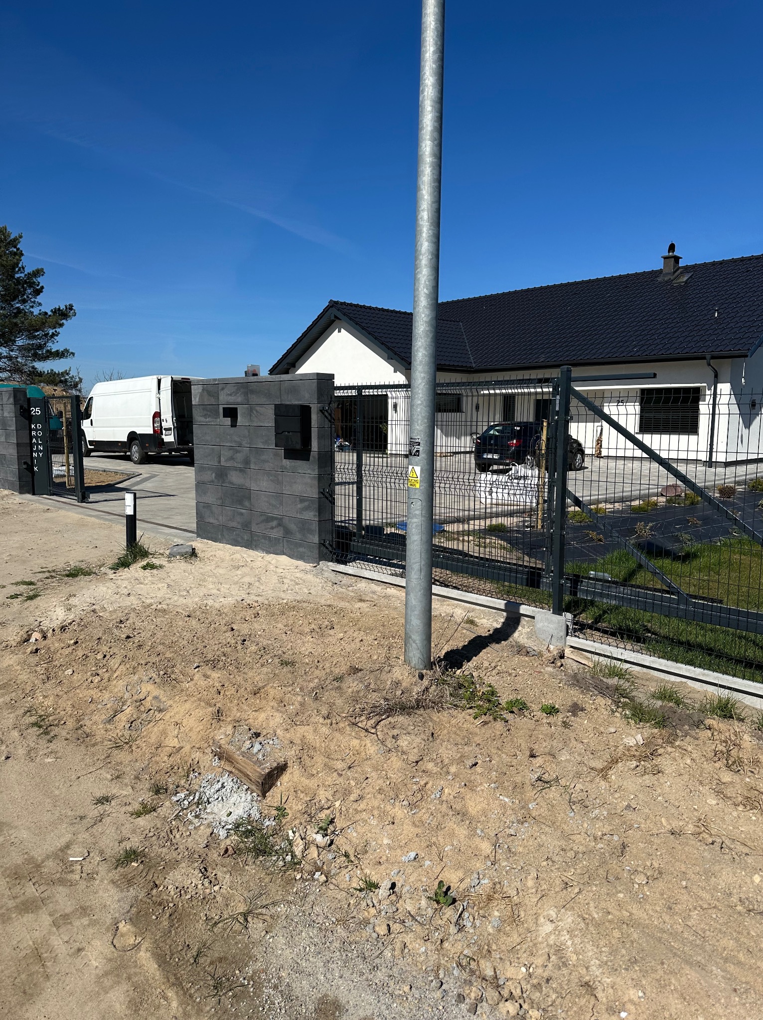 Nowoczesne ogrodzenie panelowe z bramą wjazdową na posesję z domem jednorodzinnym w słoneczny dzień. Widoczny fragment podjazdu i trawnika. Słupek oświetleniowy.