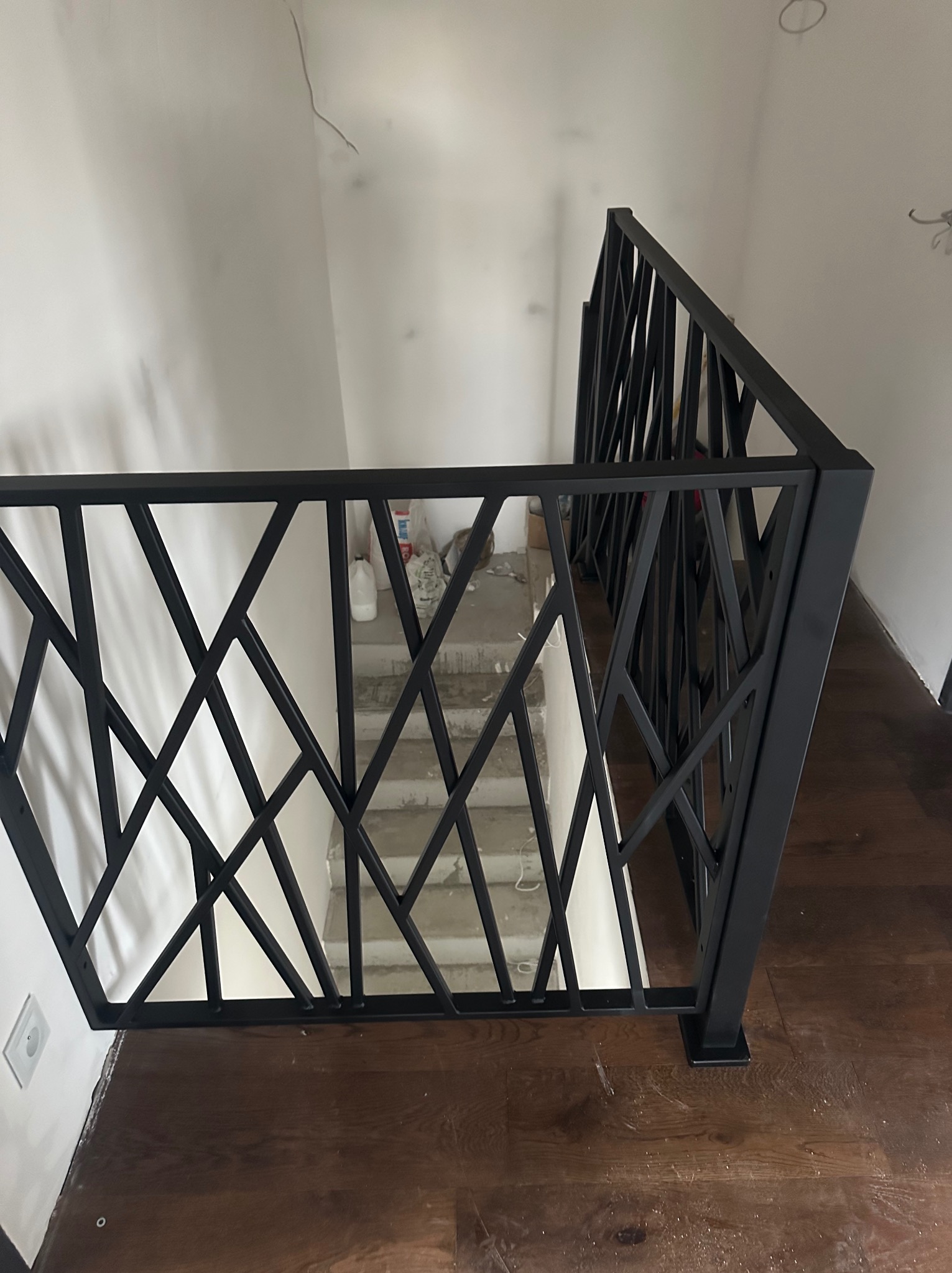 Nowoczesna, czarna balustrada metalowa o geometrycznym wzorze, zamontowana przy schodach w jasnym wnętrzu. Widoczny fragment drewnianej podłogi i surowe schody.