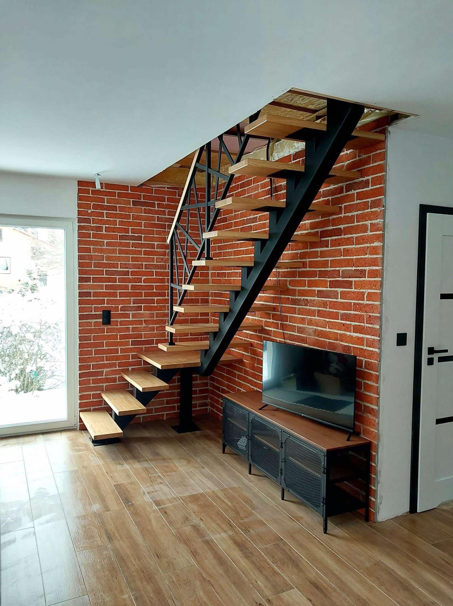 Nowoczesne schody metalowe z drewnianymi stopniami, umieszczone w pomieszczeniu z ceglaną ścianą i drewnianą podłogą. Widoczny fragment telewizora i drzwi.