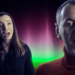 GREGOR HOME STUDIO - Dwoje śpiewających ludzi na tle cyfrowej animacji zorzy polarnej w odcieniach zieleni i różu. Kobieta z długimi włosami w ciemnej bluzce z piórami, mężczyzna w pomarańczowym golfie.