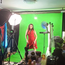 GREGOR HOME STUDIO - Kobieta w czerwonej sukience pozuje na tle zielonego ekranu w improwizowanym studiu z oświetleniem, wentylatorem i operatorem kamery, a w tle widać sofę z osobą i psem.