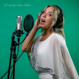 GREGOR HOME STUDIO - Wokalistka z słuchawkami śpiewa do mikrofonu z pop filtrem na tle zielonego ekranu, w studio nagrań w Opolu. Widoczny fragment napisu 'fot. Gregor Home Studio'.