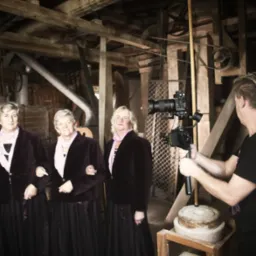 Ekipa filmowa kręci ujęcie z czterema kobietami w bordowych strojach w zabytkowym młynie, widoczne drewniane konstrukcje i kamienne elementy.