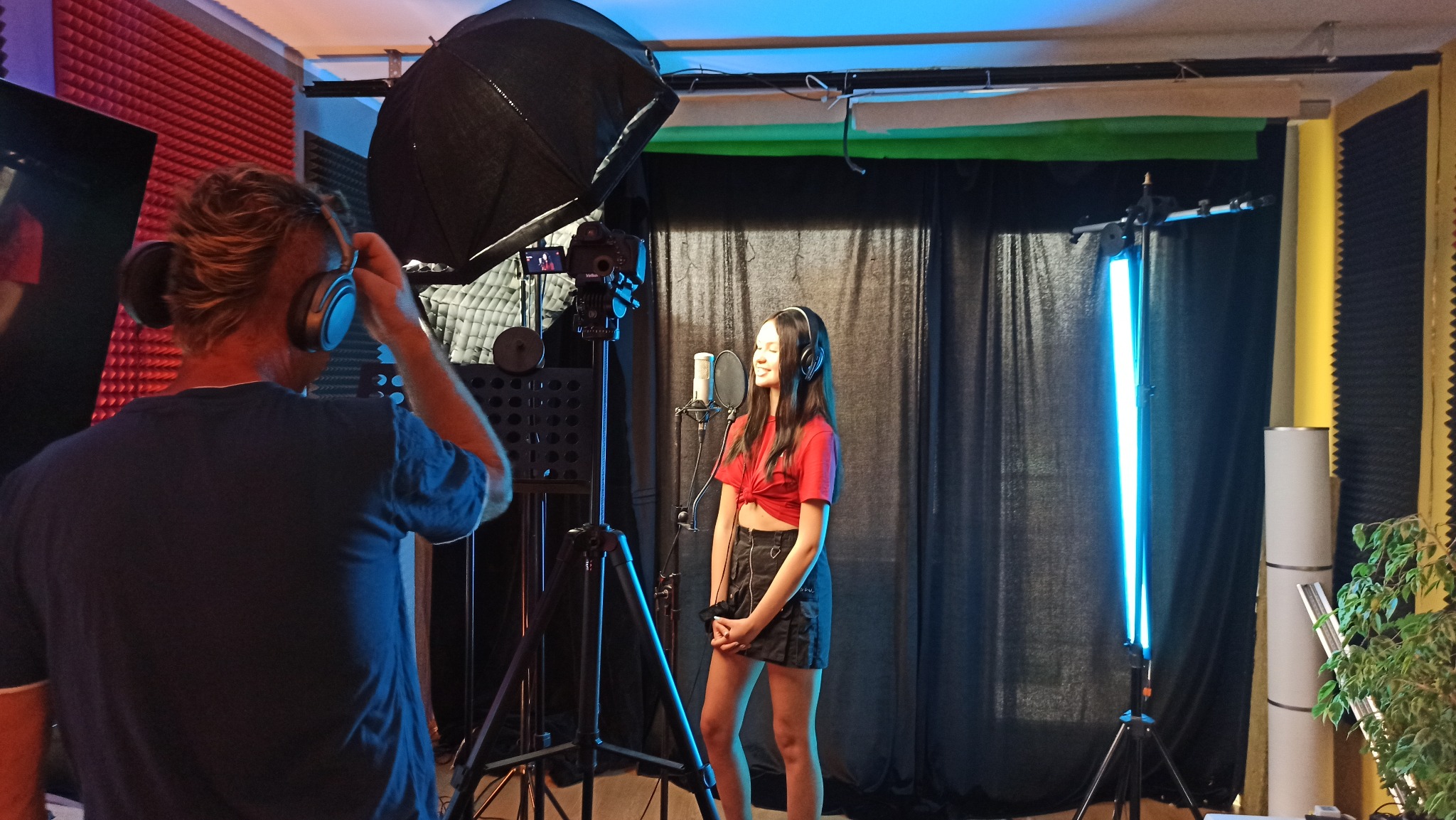 Młoda kobieta w słuchawkach śpiewa do mikrofonu w profesjonalnym studiu, oświetlona studyjnym światłem, z operatorem w tle.