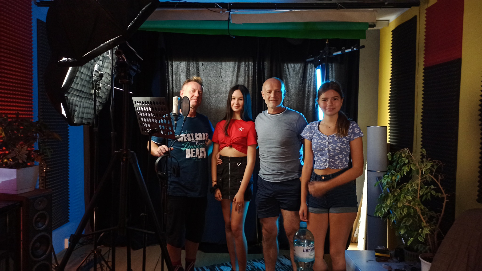 Zespół muzyczny składający się z dwóch mężczyzn i dwóch dziewczyn pozuje w profesjonalnym studiu nagraniowym z mikrofonem, statywem i oświetleniem.