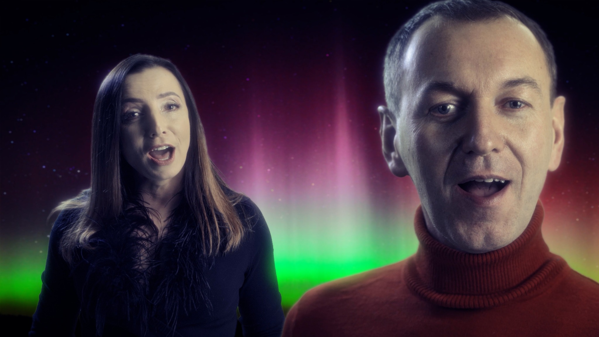 Dwoje śpiewających ludzi na tle cyfrowej animacji zorzy polarnej w odcieniach zieleni i różu. Kobieta z długimi włosami w ciemnej bluzce z piórami, mężczyzna w pomarańczowym golfie.