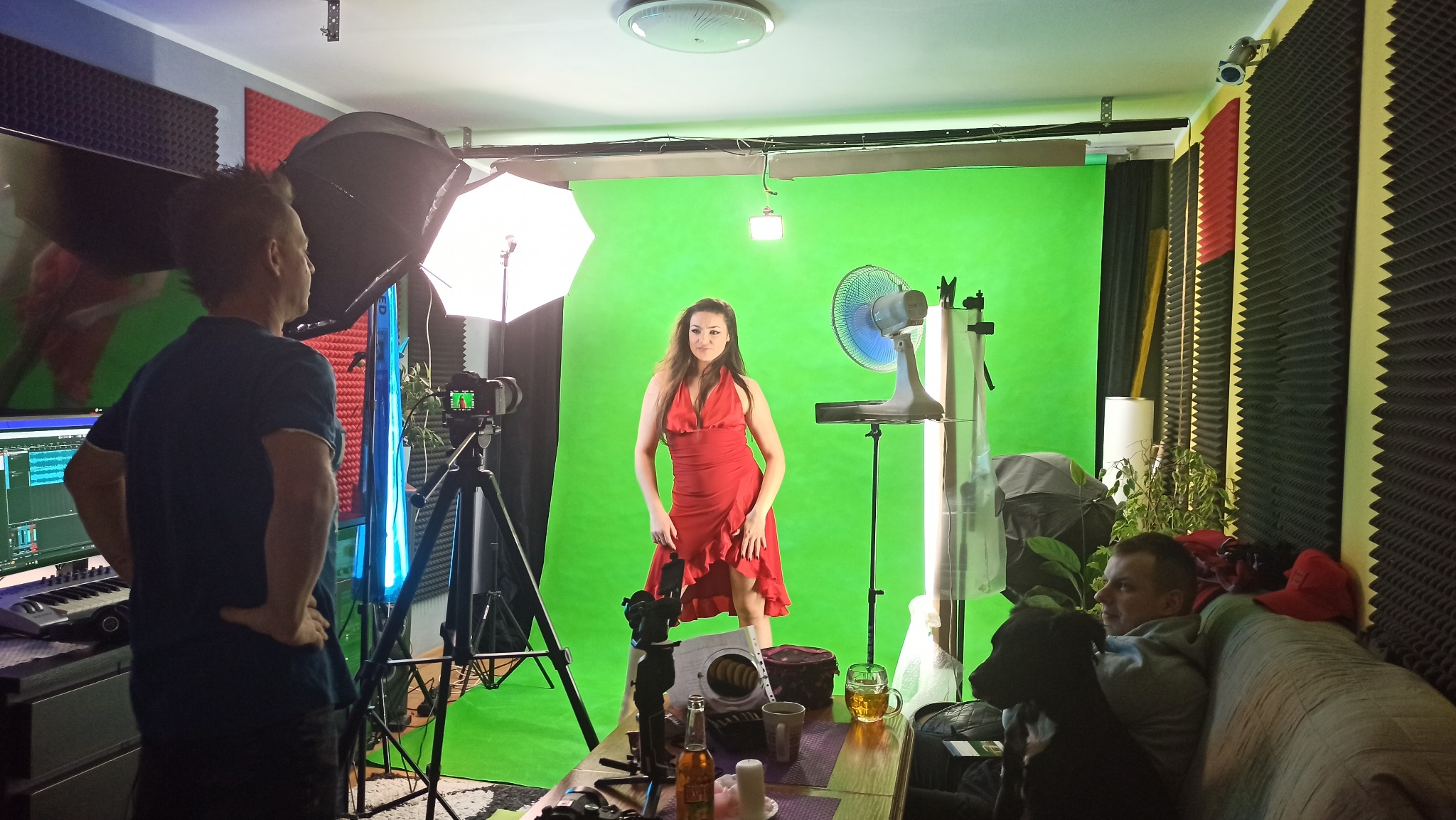 Kobieta w czerwonej sukience pozuje na tle zielonego ekranu w improwizowanym studiu z oświetleniem, wentylatorem i operatorem kamery, a w tle widać sofę z osobą i psem.