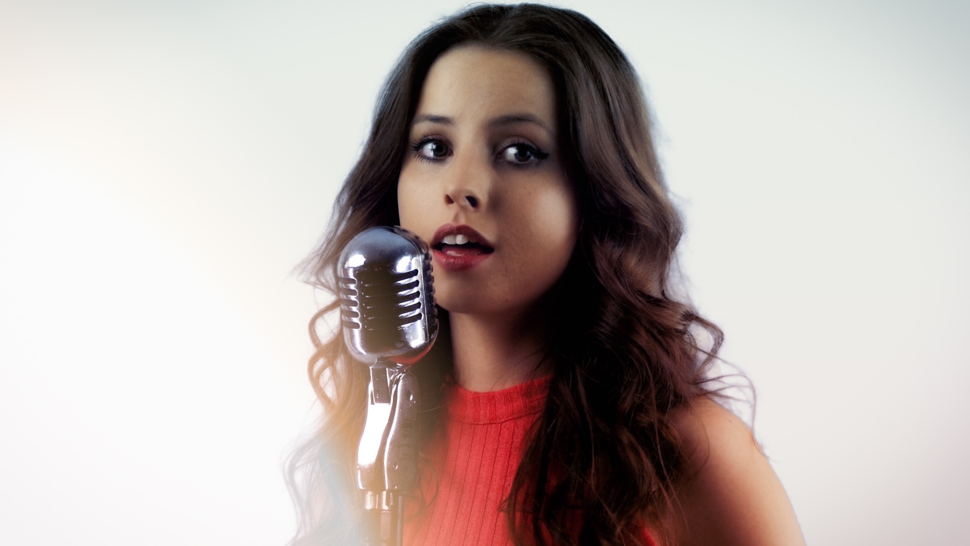 Młoda kobieta z brązowymi, falowanymi włosami śpiewa do srebrnego mikrofonu w stylu retro, z delikatnym rozświetleniem na twarzy.