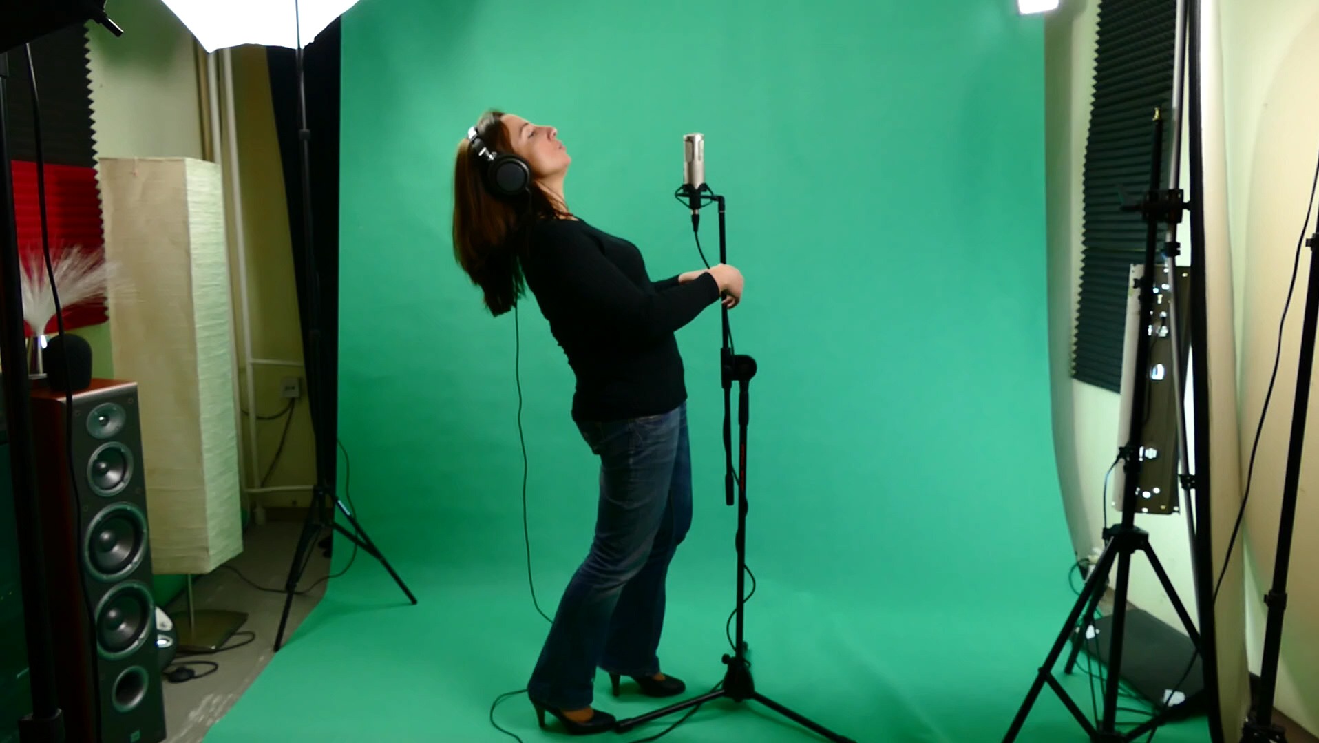 Kobieta w słuchawkach, pochylona do tyłu, śpiewa do mikrofonu w  pomieszczeniu z zielonym tłem, widoczne oświetlenie studyjne i kolumna głośnikowa.
