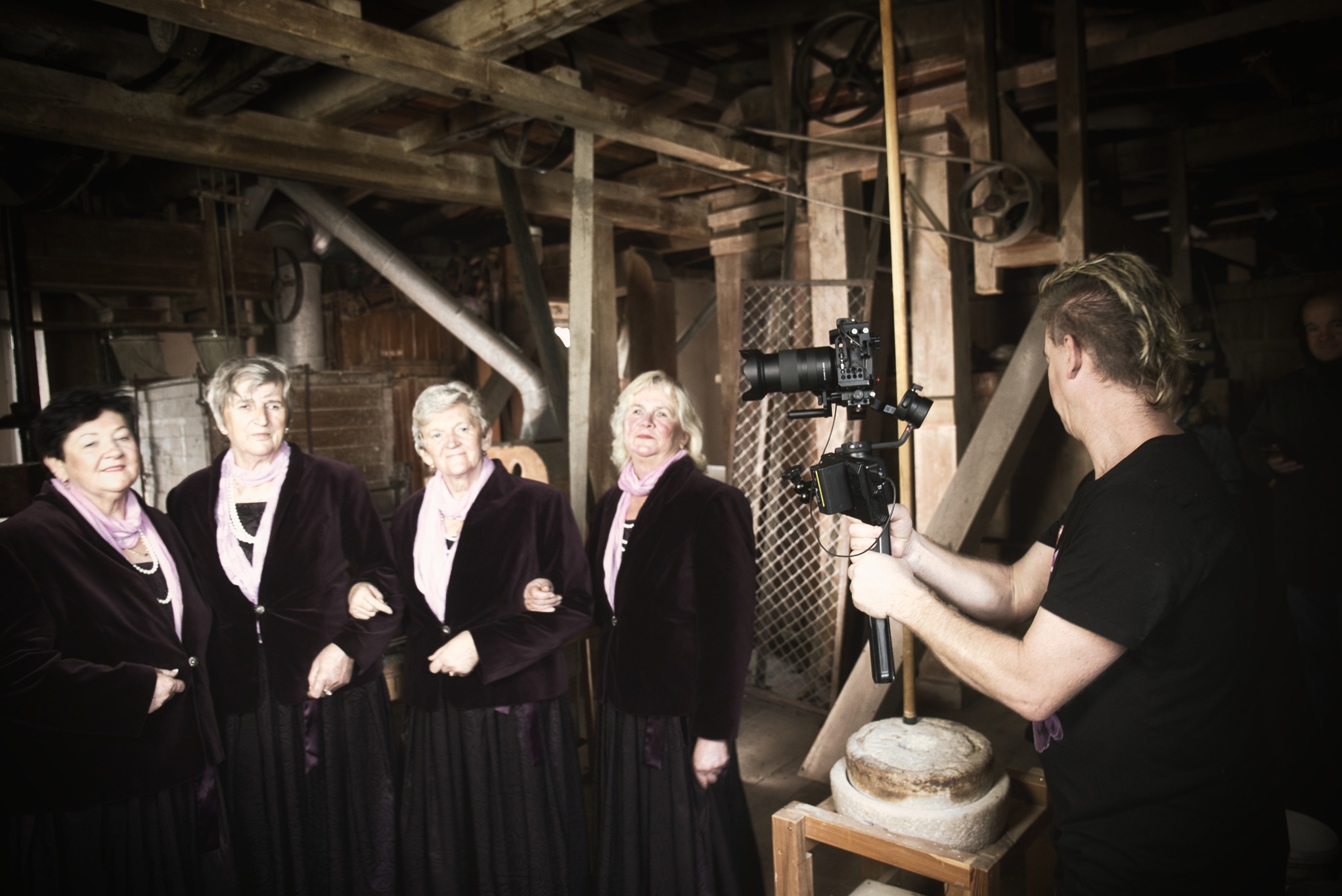 Ekipa filmowa kręci ujęcie z czterema kobietami w bordowych strojach w zabytkowym młynie, widoczne drewniane konstrukcje i kamienne elementy.