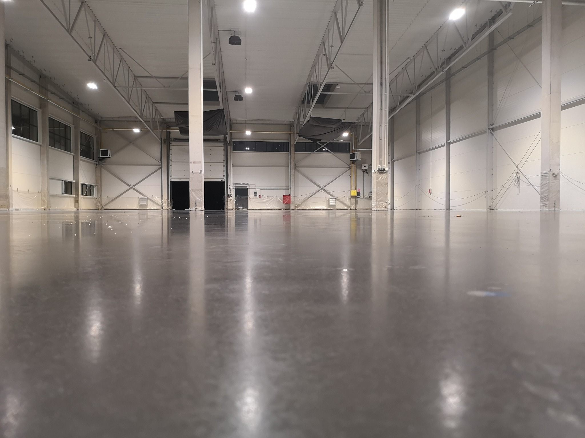 Polerowana, szara posadzka przemysłowa w hali magazynowej z widocznymi stalowymi słupami i bramami w tle.