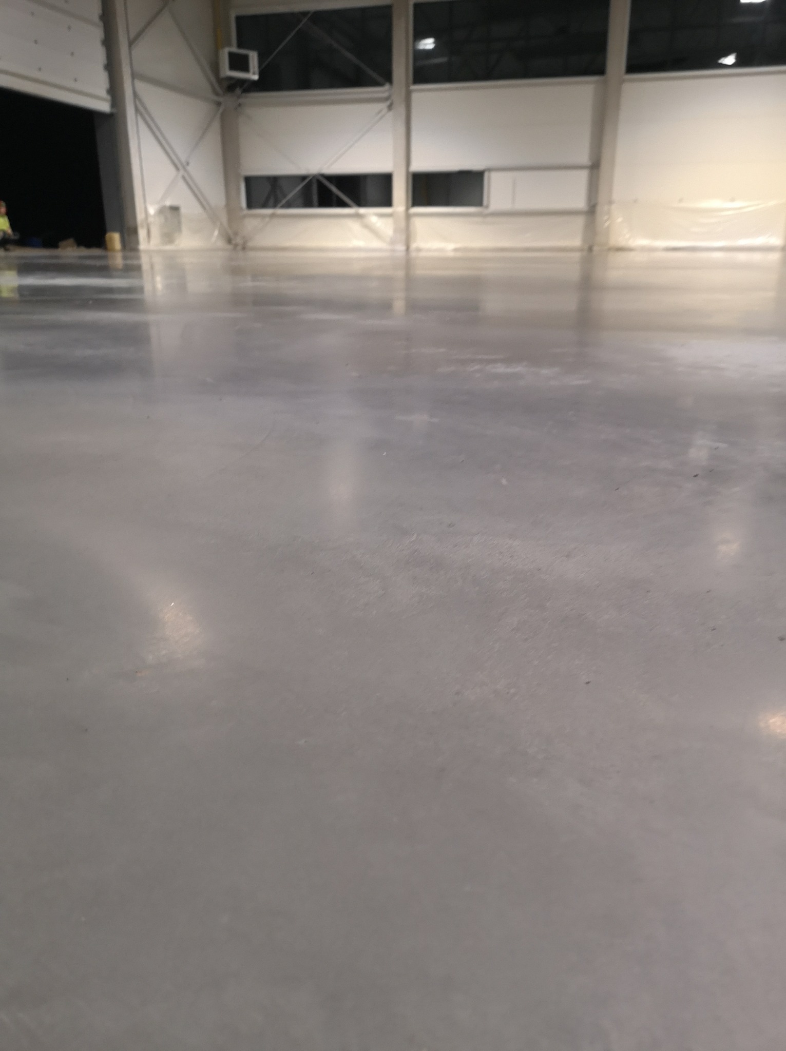 Błyszcząca, szara posadzka przemysłowa w hali produkcyjnej, widoczne refleksy światła, w tle fragment konstrukcji hali i otwarte wrota.
