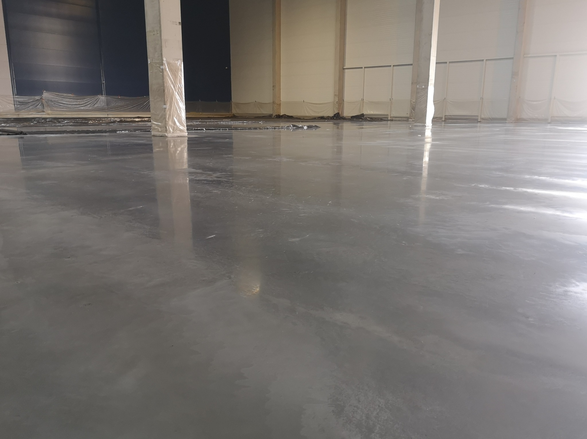 Świeżo wylana, gładka, szara posadzka w hali przemysłowej, z zabezpieczonymi folią słupami nośnymi w tle.
