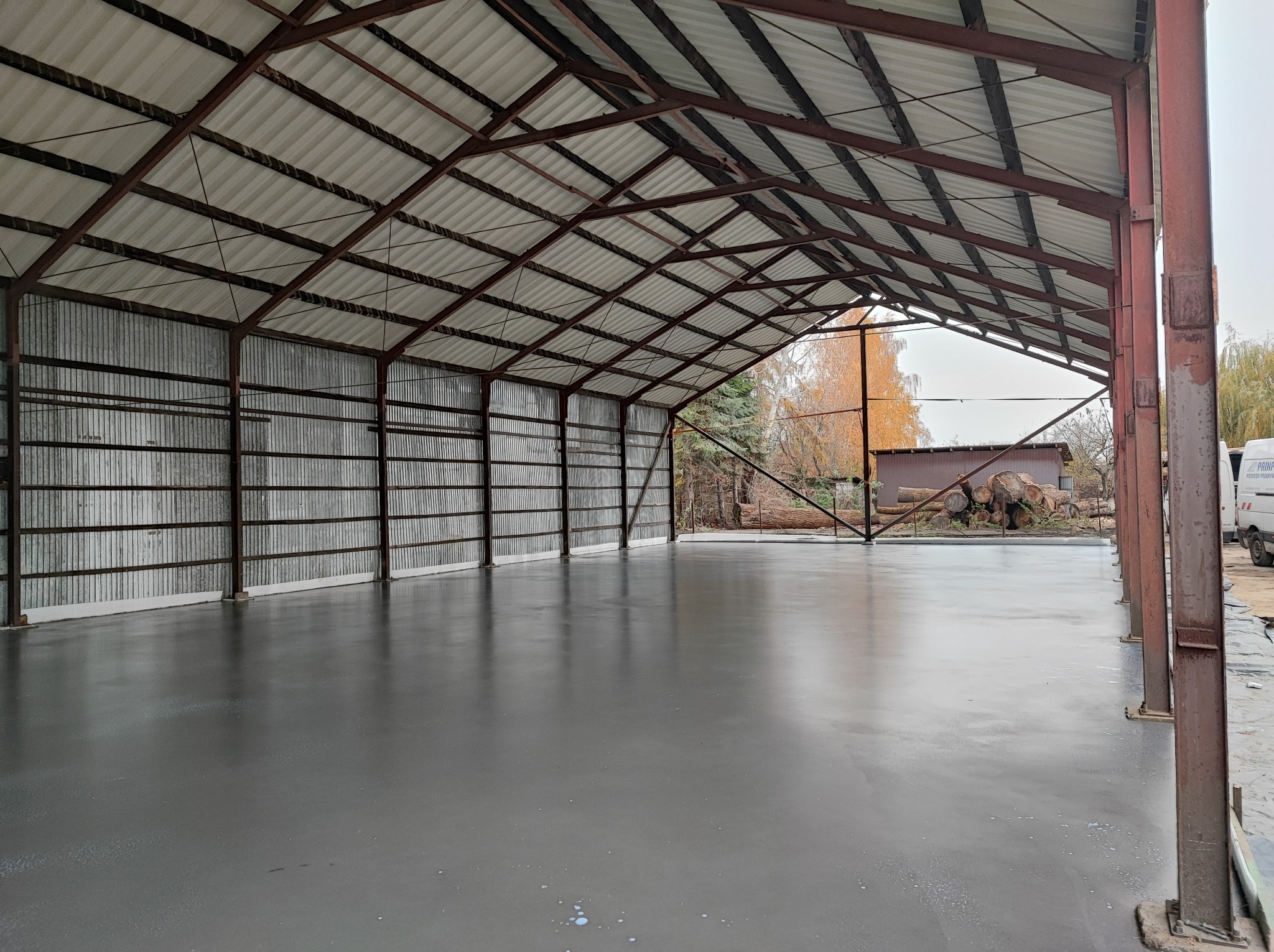 Świeżo wylana, gładka, szara posadzka betonowa wewnątrz hali magazynowej o konstrukcji stalowej, widoczne krople wody na powierzchni betonu, drzewa w tle.