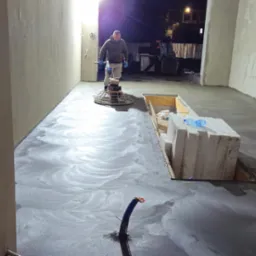 Pracownik w szarej bluzie wygładza wylewkę jastrychową za pomocą elektrycznej zacieraczki do betonu w surowym wnętrzu, widoczne białe bloczki i butelka wody.