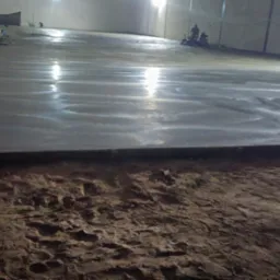 Wnętrze hali produkcyjnej w trakcie wylewania i wygładzania posadzki, widoczne maszyny do wygładzania i dwóch pracowników, w tle ściany hali, pierwszy plan z fragmentem podłoża przed wylewką.