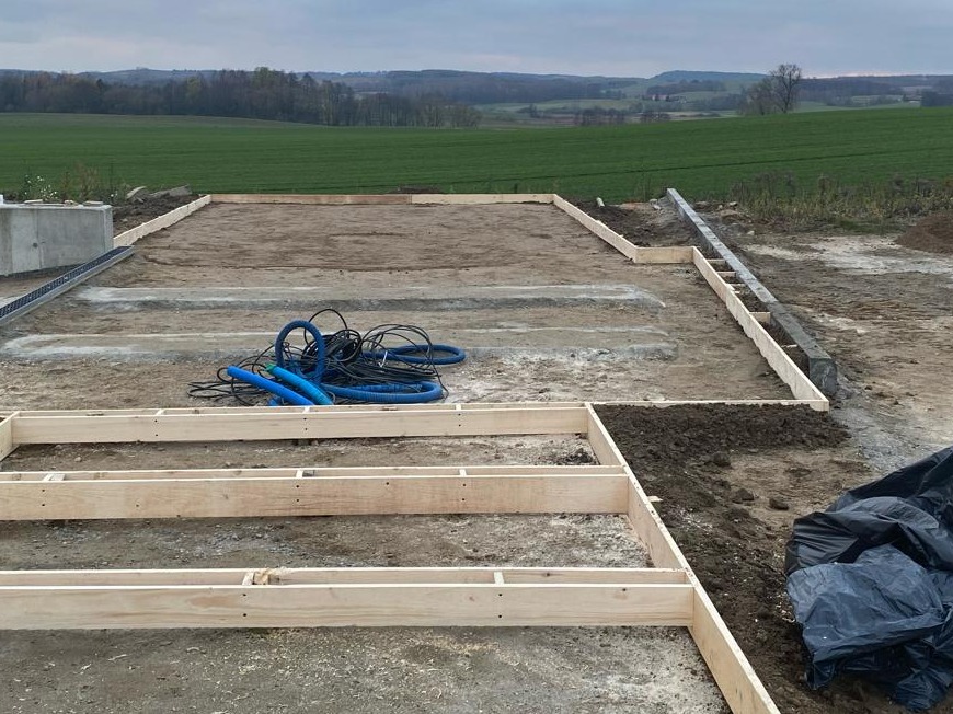 Przygotowanie podłoża pod taras: drewniana konstrukcja szkieletowa z legarów, widoczne rury instalacyjne i betonowy element odwodnienia na tle krajobrazu polnych wzgórz.