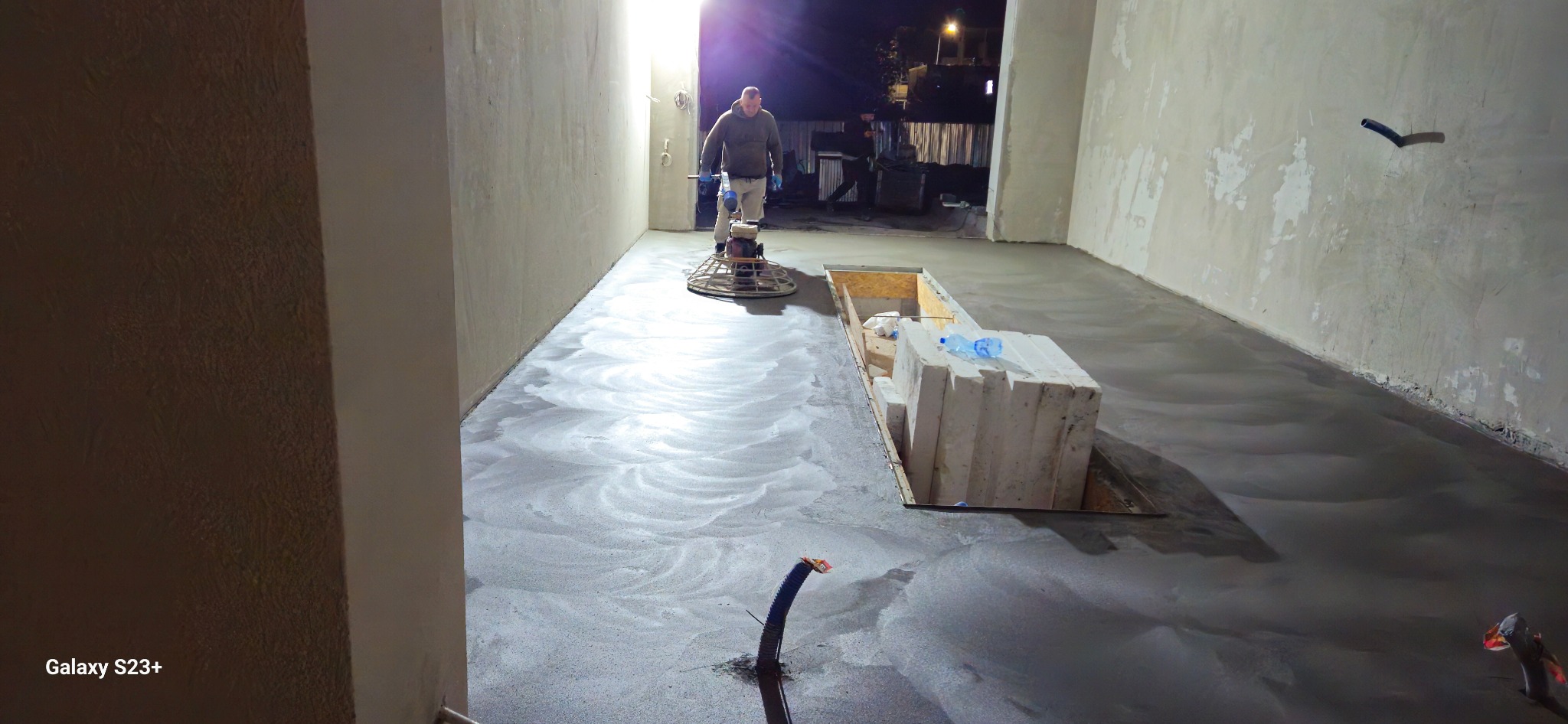 Pracownik w szarej bluzie wygładza wylewkę jastrychową za pomocą elektrycznej zacieraczki do betonu w surowym wnętrzu, widoczne białe bloczki i butelka wody.