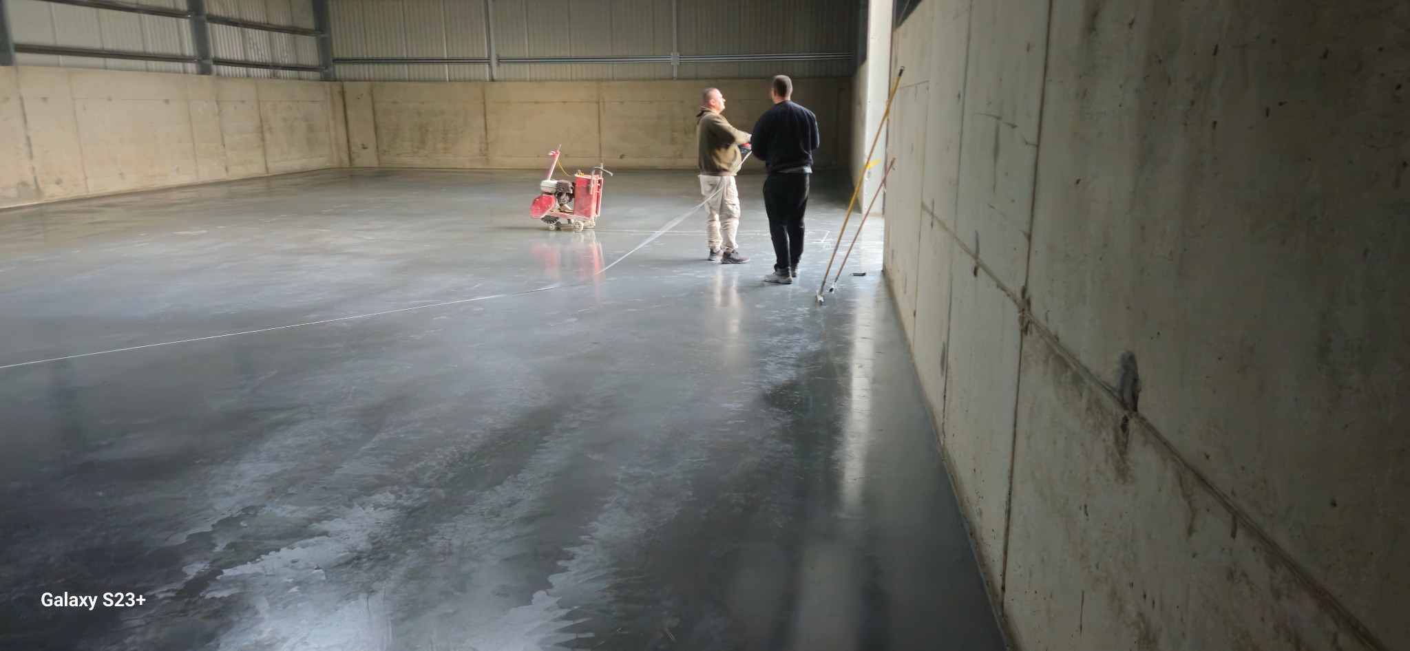 Wykończona, błyszcząca posadzka w hali przemysłowej, z widocznymi dwoma pracownikami i maszyną do szlifowania w tle, oświetlenie naturalne z góry.