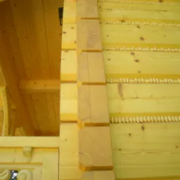 Fragment drewnianej konstrukcji domu z bali, widoczne połączenia bali z użyciem drewnianych kołków, detale ciesielskie i wykończenie elewacji z dekoracyjnymi elementami.