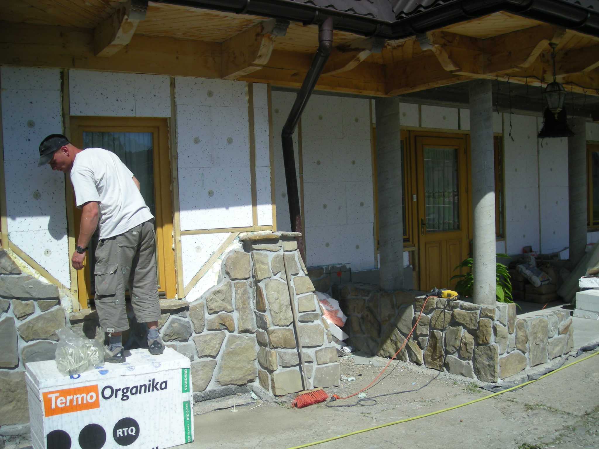 Pracownik w trakcie montażu izolacji elewacyjnej ze styropianu, stojący na opakowaniu materiału termoizolacyjnego, widoczne fragmenty elewacji wykończone kamieniem naturalnym, drewniane elementy...
