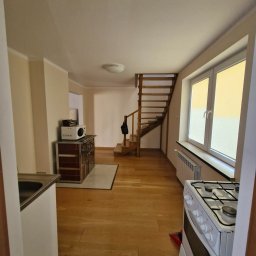 Ilona Zawadzka - Jasne wnętrze domu po remoncie: drewniana podłoga, schody, okno z roletami, piec kaflowy z mikrofalówką, biała kuchenka. Widok z perspektywy wejścia.