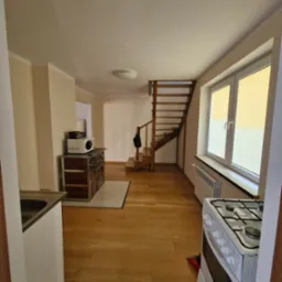 Jasne wnętrze domu po remoncie: drewniana podłoga, schody, okno z roletami, piec kaflowy z mikrofalówką, biała kuchenka. Widok z perspektywy wejścia.