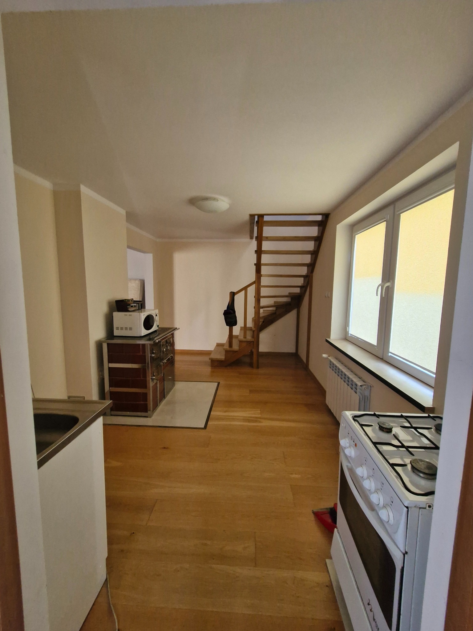 Jasne wnętrze domu po remoncie: drewniana podłoga, schody, okno z roletami, piec kaflowy z mikrofalówką, biała kuchenka. Widok z perspektywy wejścia.