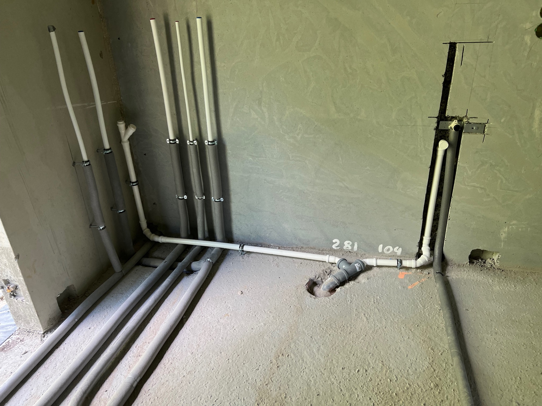 Surowe wnętrze z widocznymi rurami instalacji wodnej i kanalizacyjnej w trakcie montażu, pionowe rury zabezpieczone izolacją, poziome rury z szarymi złączkami i oznaczeniami numerów na ścianie.
