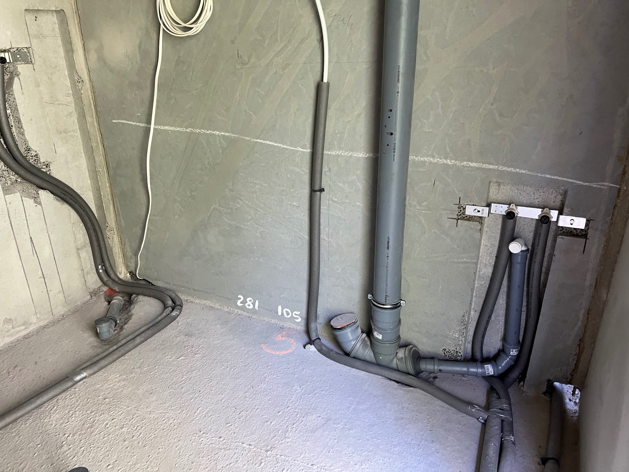 Surowe wnętrze łazienki w trakcie instalacji: szare rury kanalizacyjne i grzewcze prowadzone po podłodze i ścianach, widoczne oznaczenia na podłodze, przygotowane punkty podłączeń.