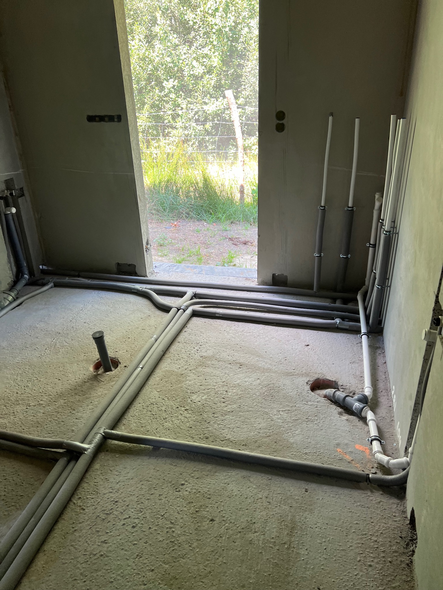 Szare rury instalacji wodnej i kanalizacyjnej ułożone na surowym betonowym podłożu w trakcie montażu, widoczne otwory w podłodze i wyprowadzone piony przy ścianie, surowe ściany w tle.