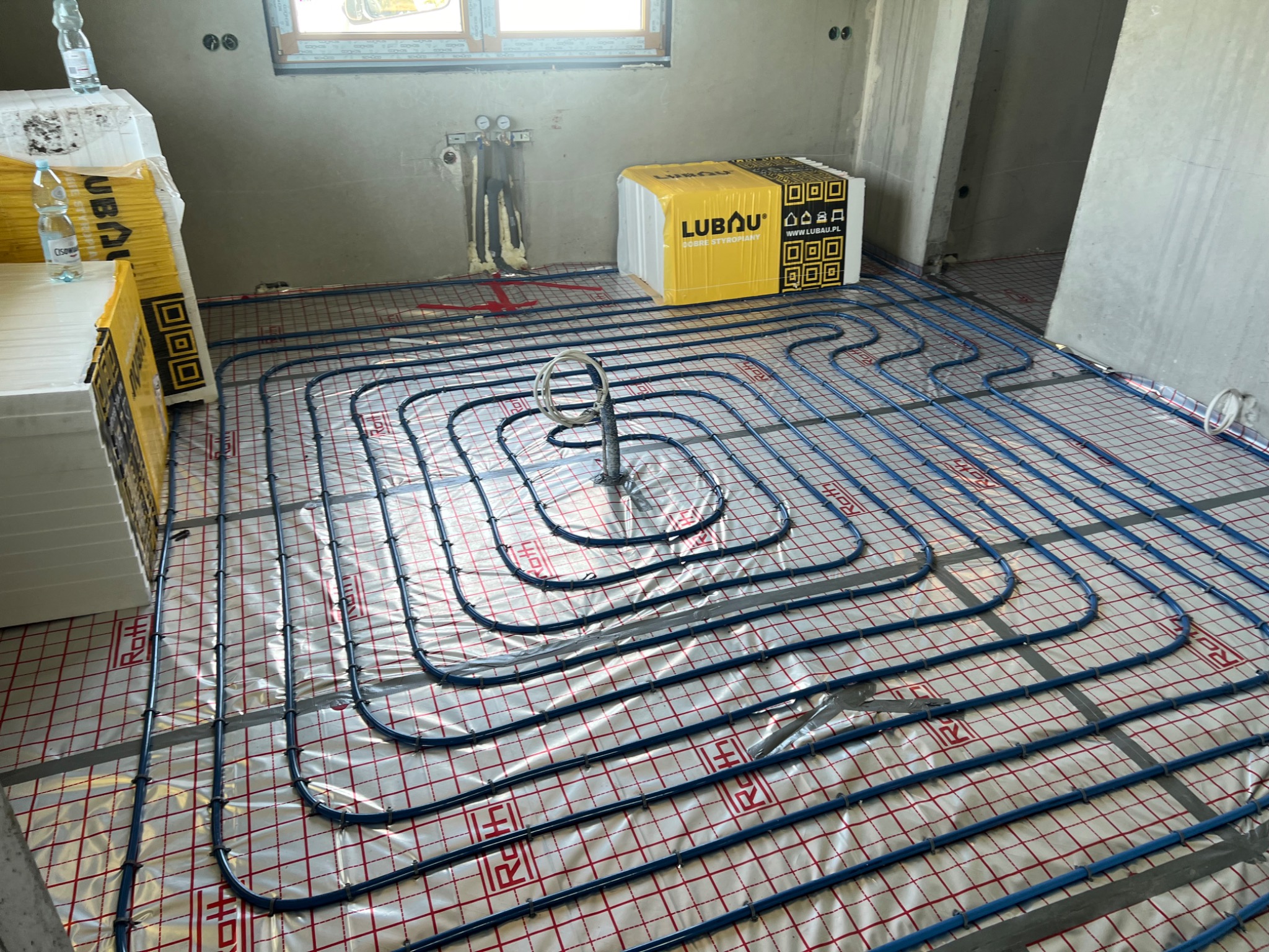 Niebieskie rury ogrzewania podłogowego ułożone spiralnie na folii z kratką, widoczne materiały izolacyjne i pakiet styropianu Lubau w surowym wnętrzu budynku.