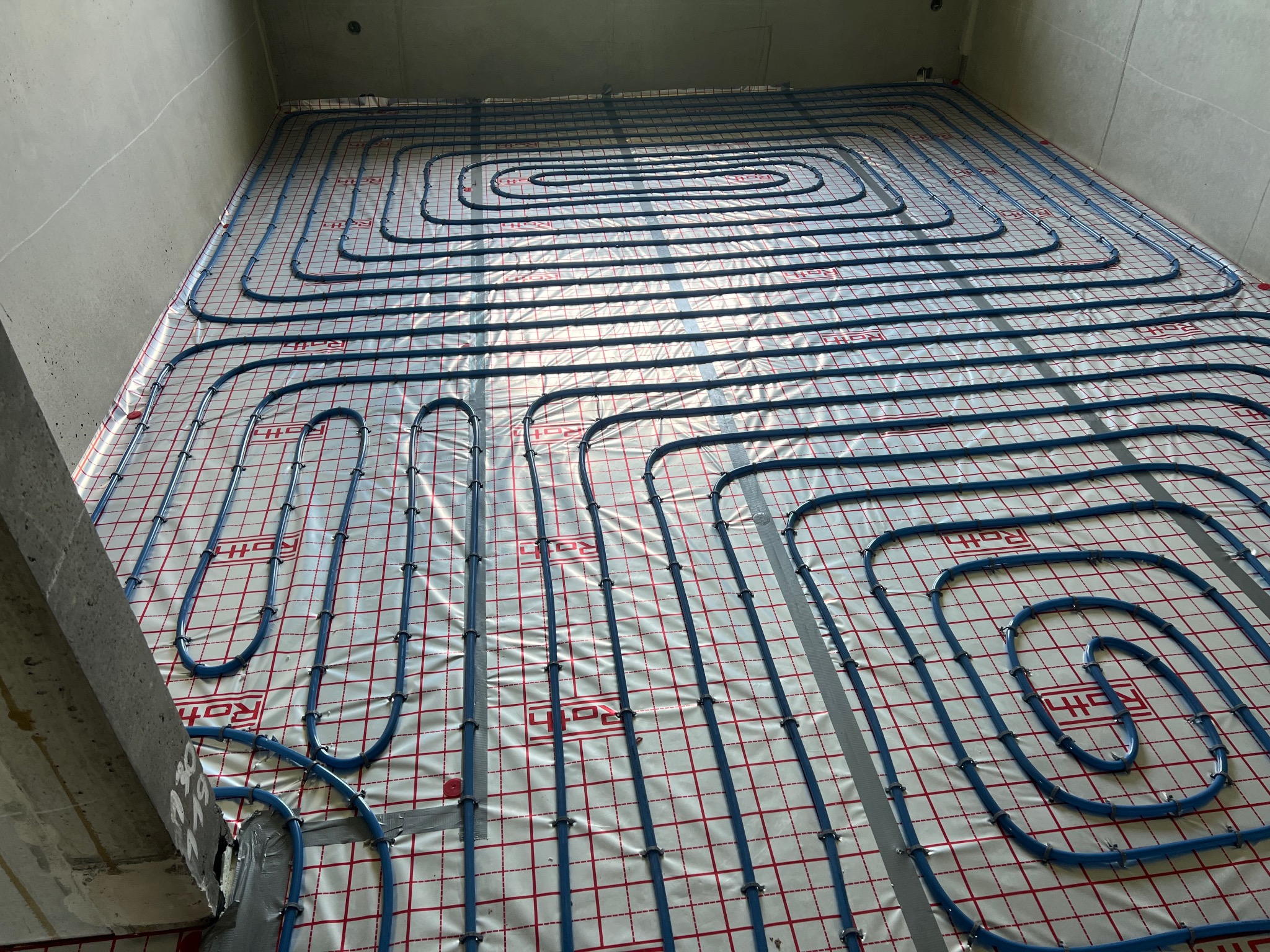 Niebieskie rury ogrzewania podłogowego ułożone spiralnie na folii z czerwoną kratką, przymocowane taśmą i plastikowymi klipsami, widok z góry w surowym wnętrzu budynku.
