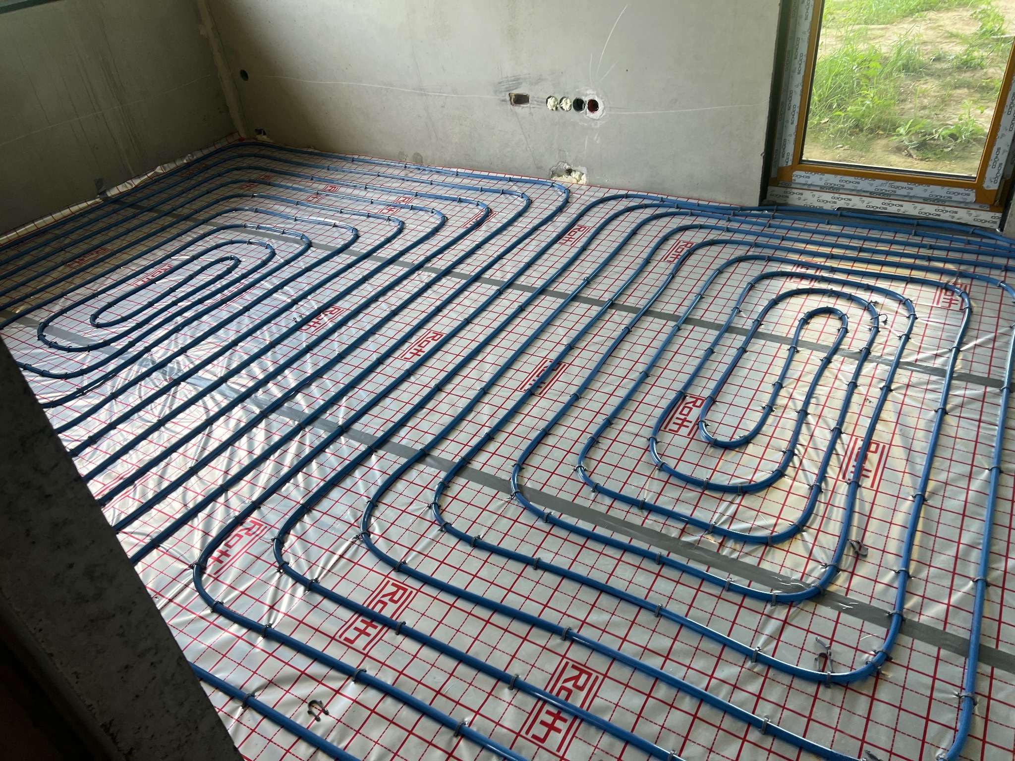 Niebieskie rury ogrzewania podłogowego ułożone na folii z siatką w surowym pomieszczeniu z oknem, widoczne otwory w ścianie.