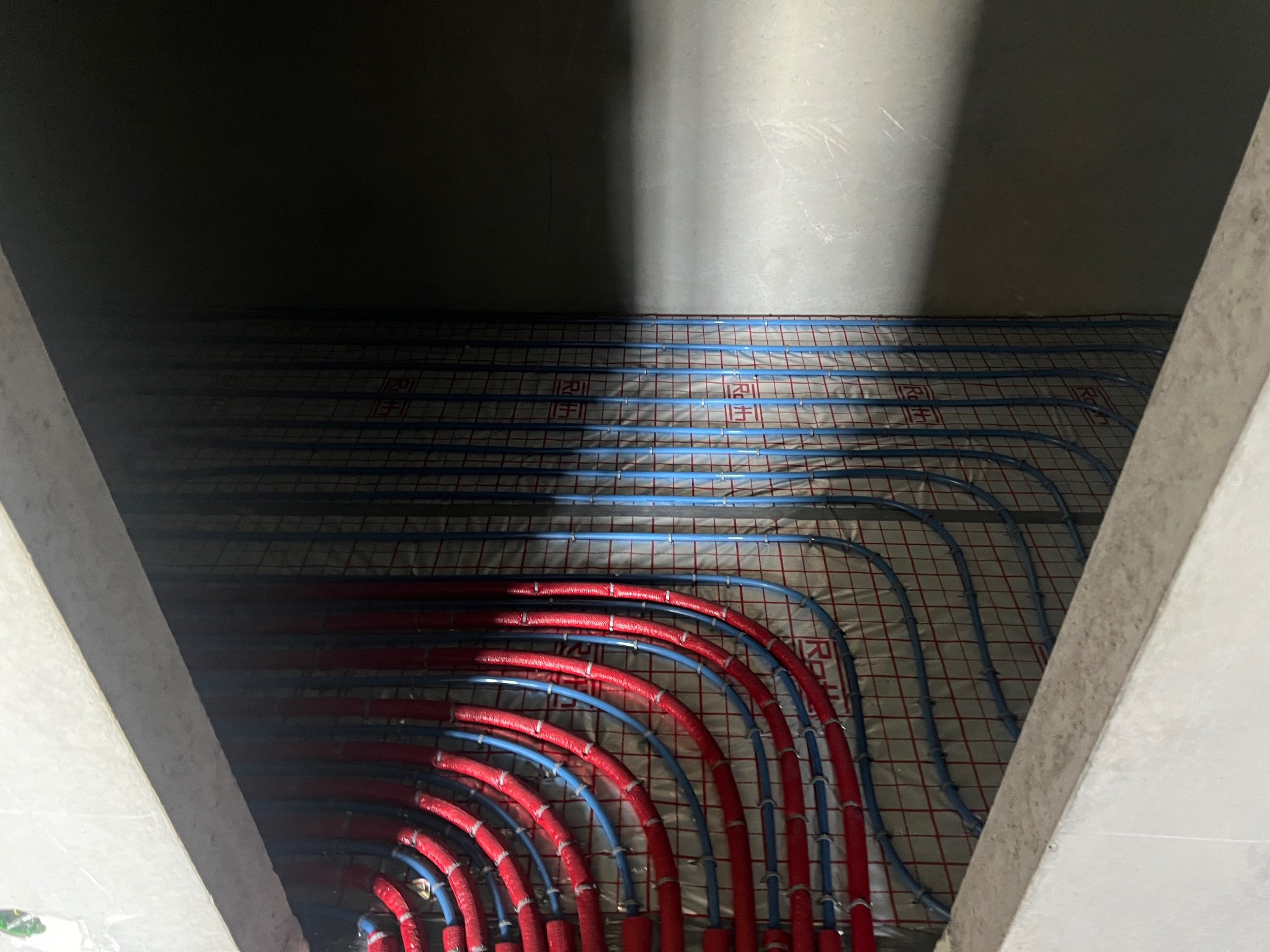 Niedokończona instalacja ogrzewania podłogowego z rurami w kolorze czerwonym i niebieskim na podkładzie z siatką, widok z dołu w surowym wnętrzu.