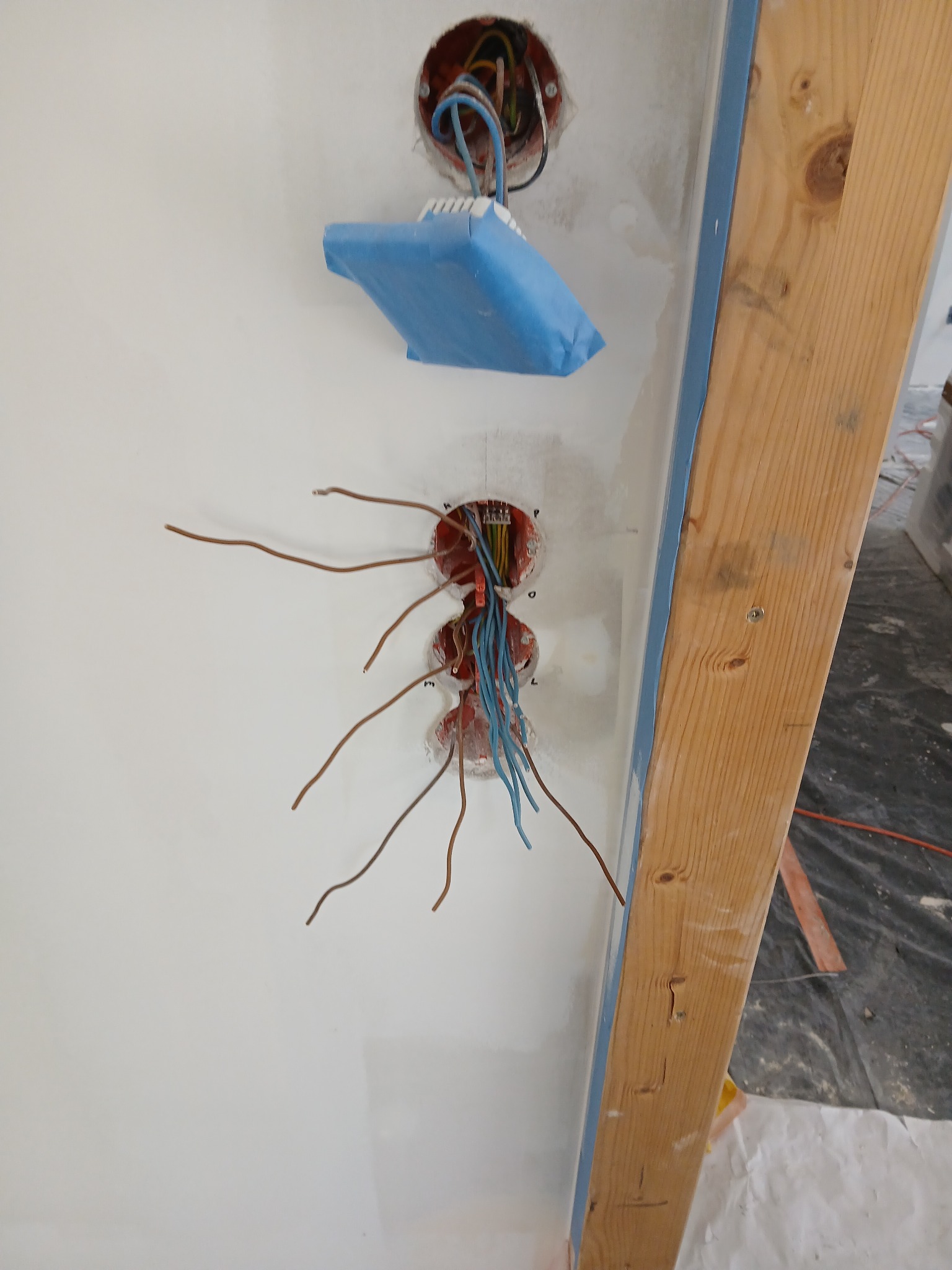 Ściana z trzema puszkami elektrycznymi, z których wystają przewody. Jedna puszka zabezpieczona niebieską taśmą. Widoczny fragment drewnianej ramy drzwi.