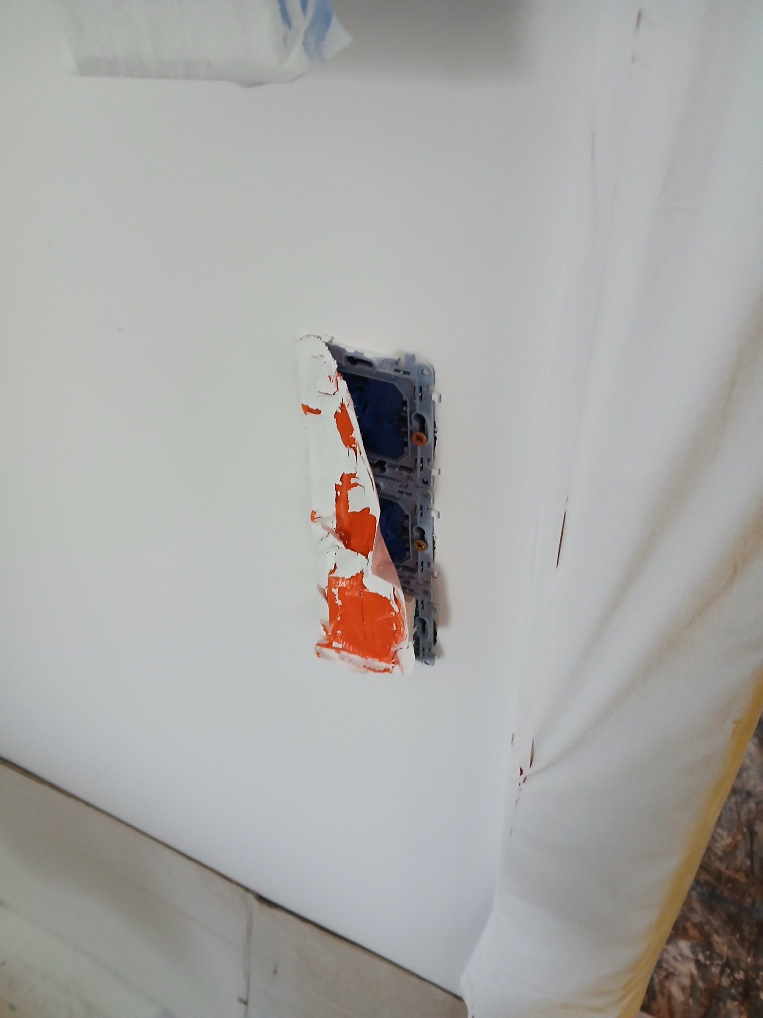 Gniazdko elektryczne w ścianie, zabezpieczone taśmą malarską z fragmentami farby, widoczne ślady prac malarskich na ścianie i folia ochronna.