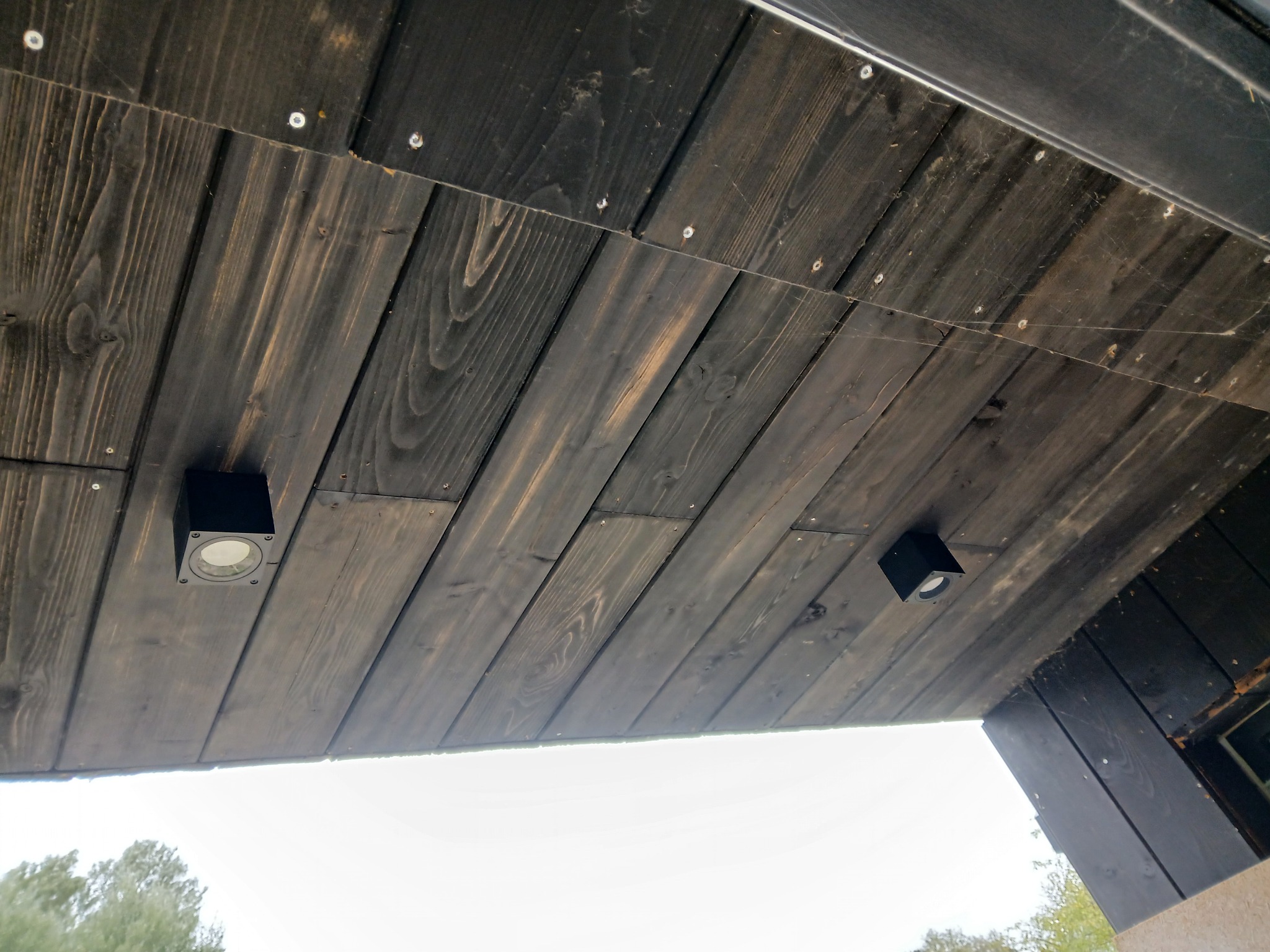 Dwa czarne reflektory punktowe zamontowane na drewnianym, ciemnym suficie podwieszanym z widocznymi śrubami mocującymi. Ujęcie z dołu.