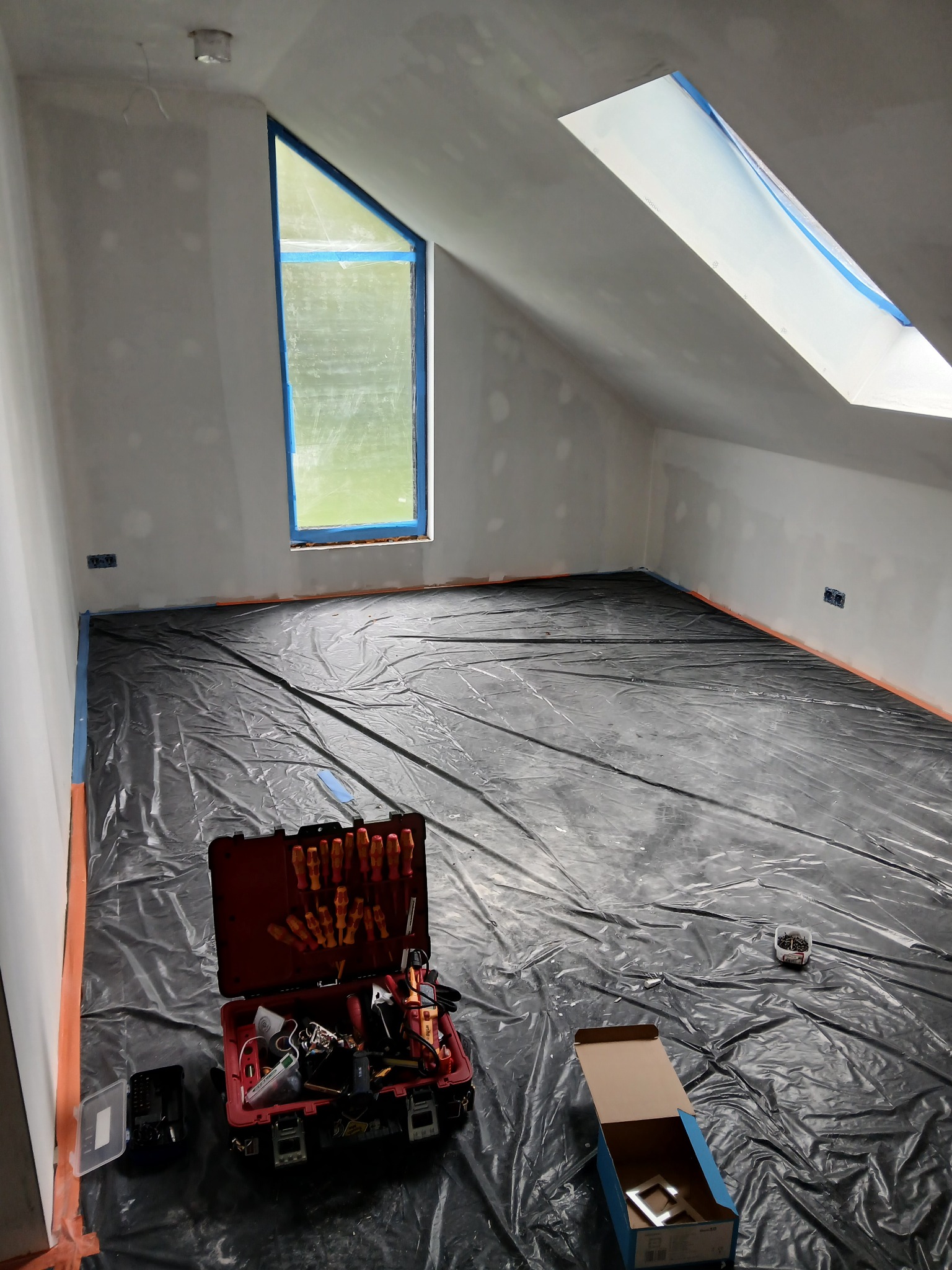 Pokój na poddaszu przygotowany do prac elektrycznych: zabezpieczona folią podłoga, okna i ściany, skrzynka z narzędziami elektrycznymi na pierwszym planie.
